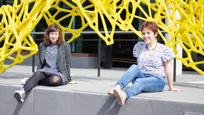 Two PhD students sitting on a concrete block in front of a yellow metal sculpture