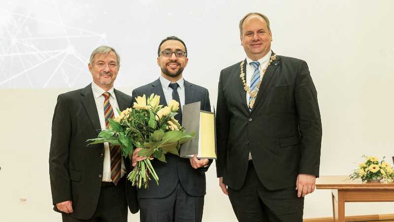 Laureate Nicola Mitwasi holding his certificate and a bouquet of flowers, with Prof. Michael Bachmann (left) and Dresden’s Lord Mayor Dirk Hilbert (right), all looking into the camera